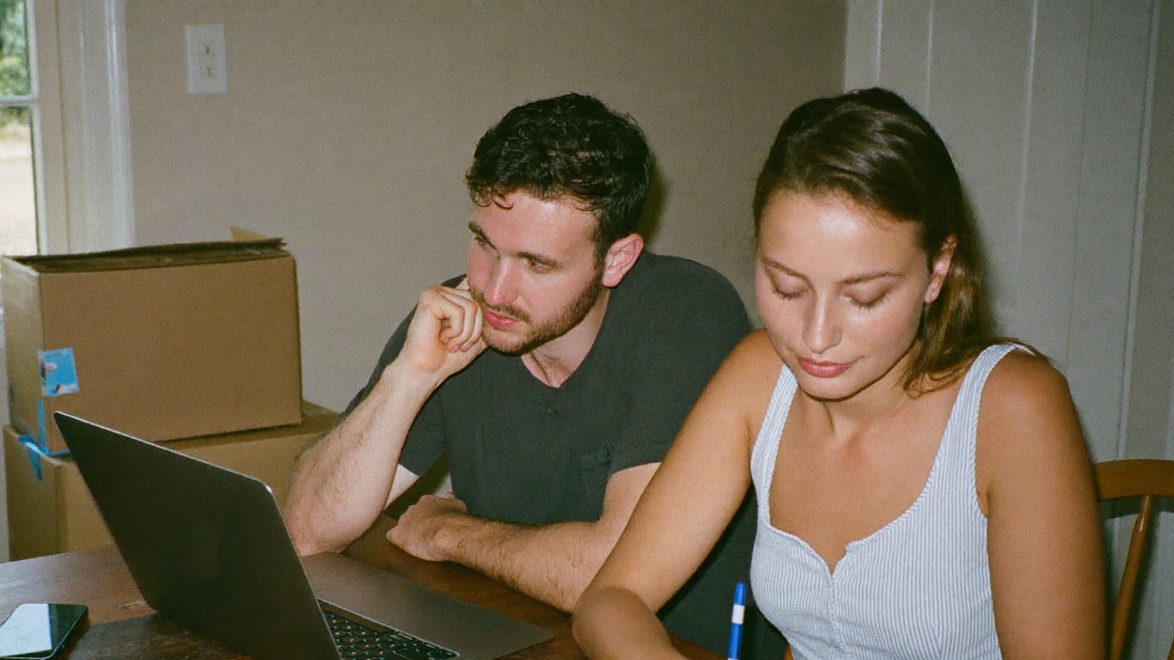 Two people at a kitchen table planning a move on a laptop, with moving boxes behind them